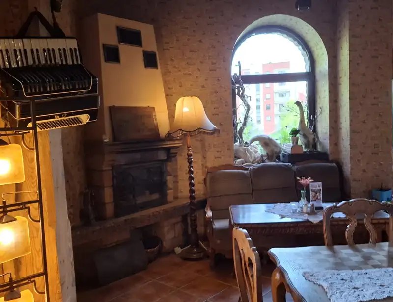 Kącik przy kominku: fotel, lampa stojąca, akordeon na ceglanej ścianie i łukowe okno